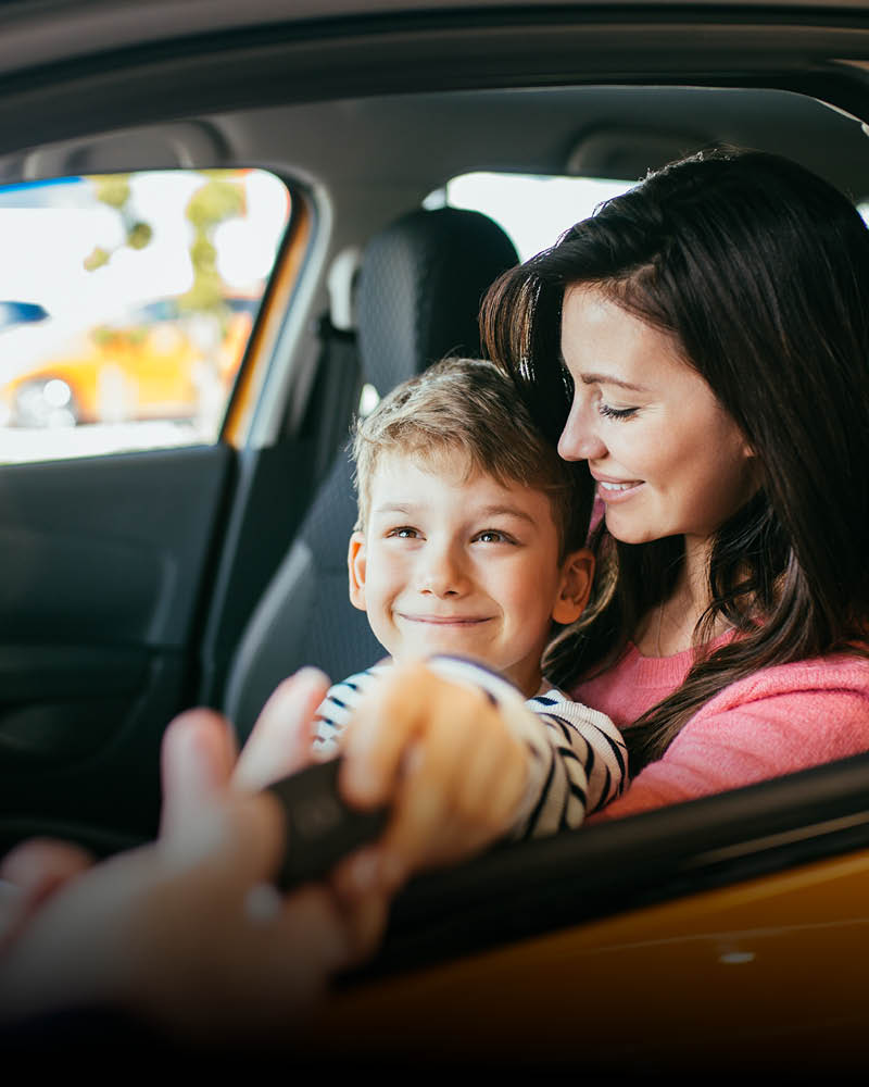 Eine Frau sitzt mit einem Kind auf dem Beifahrersitz eines Autos und nimmt einen Autoschlüssel entgegen.