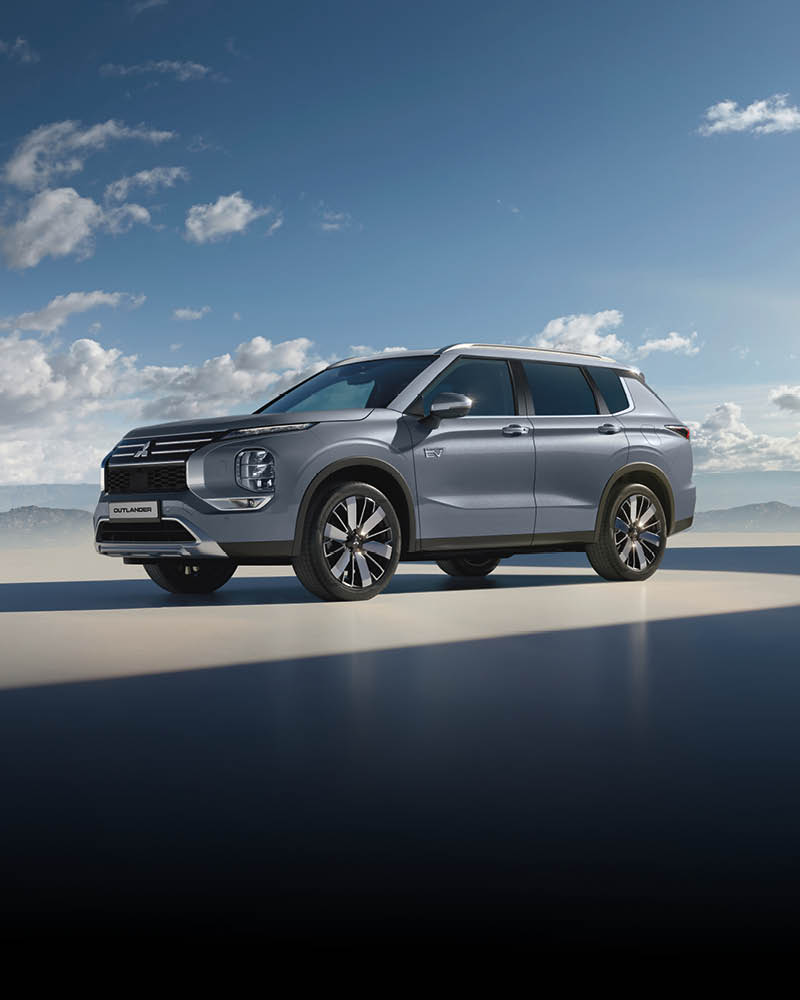 Das Bild zeigt einen silbernen SUV (Mitsubishi Outlander) auf einer ebenen Fläche unter blauem Himmel mit einigen Wolken.