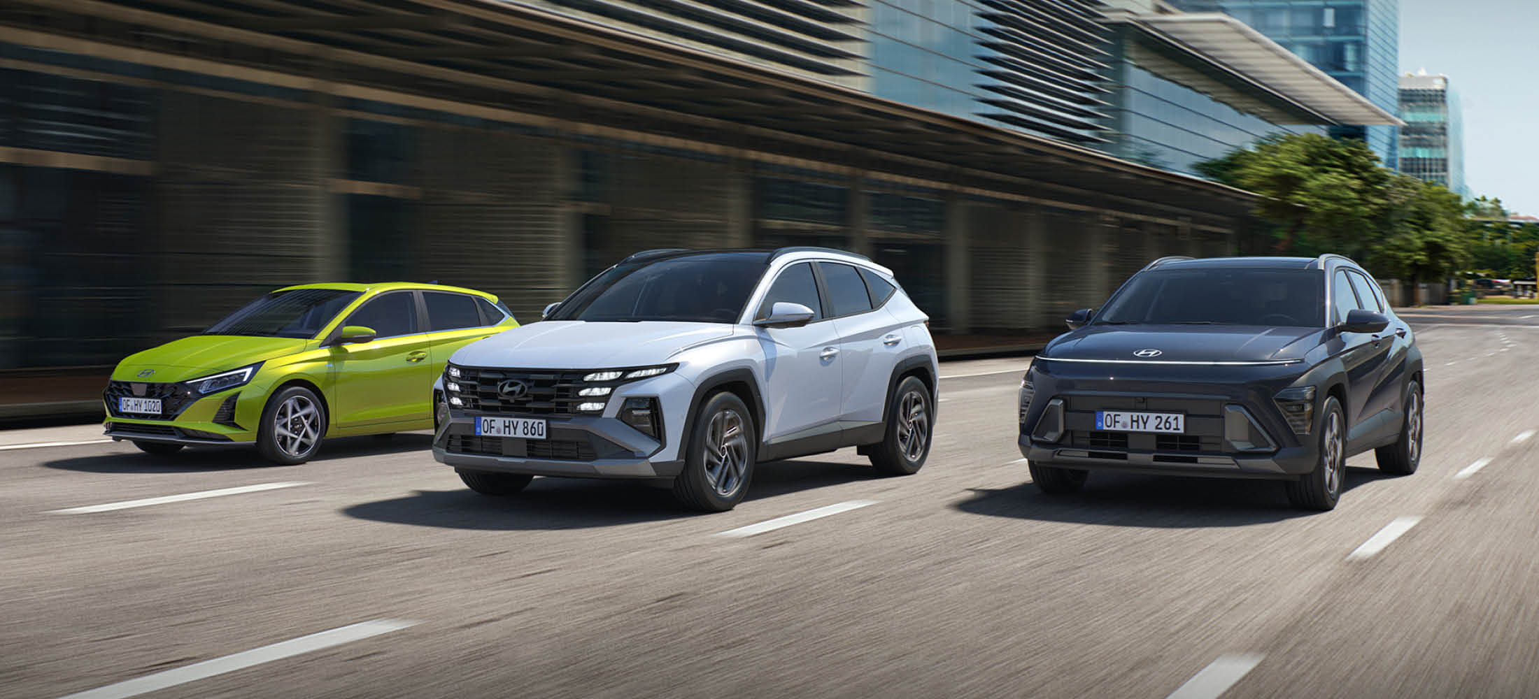 Drei moderne Autos fahren nebeneinander auf einer mehrspurigen Straße in der Stadt. Die Fahrzeuge sind unterschiedlich lackiert – eines leuchtend grün, eines weiß und eines dunkelgrau.