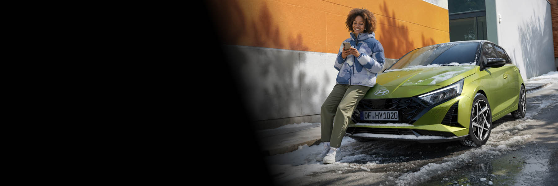 Eine Person lehnt entspannt mit ihrem Smartphone an einem grünen Hyundai, der in einer winterlichen Umgebung vor einem modernen Gebäude steht.