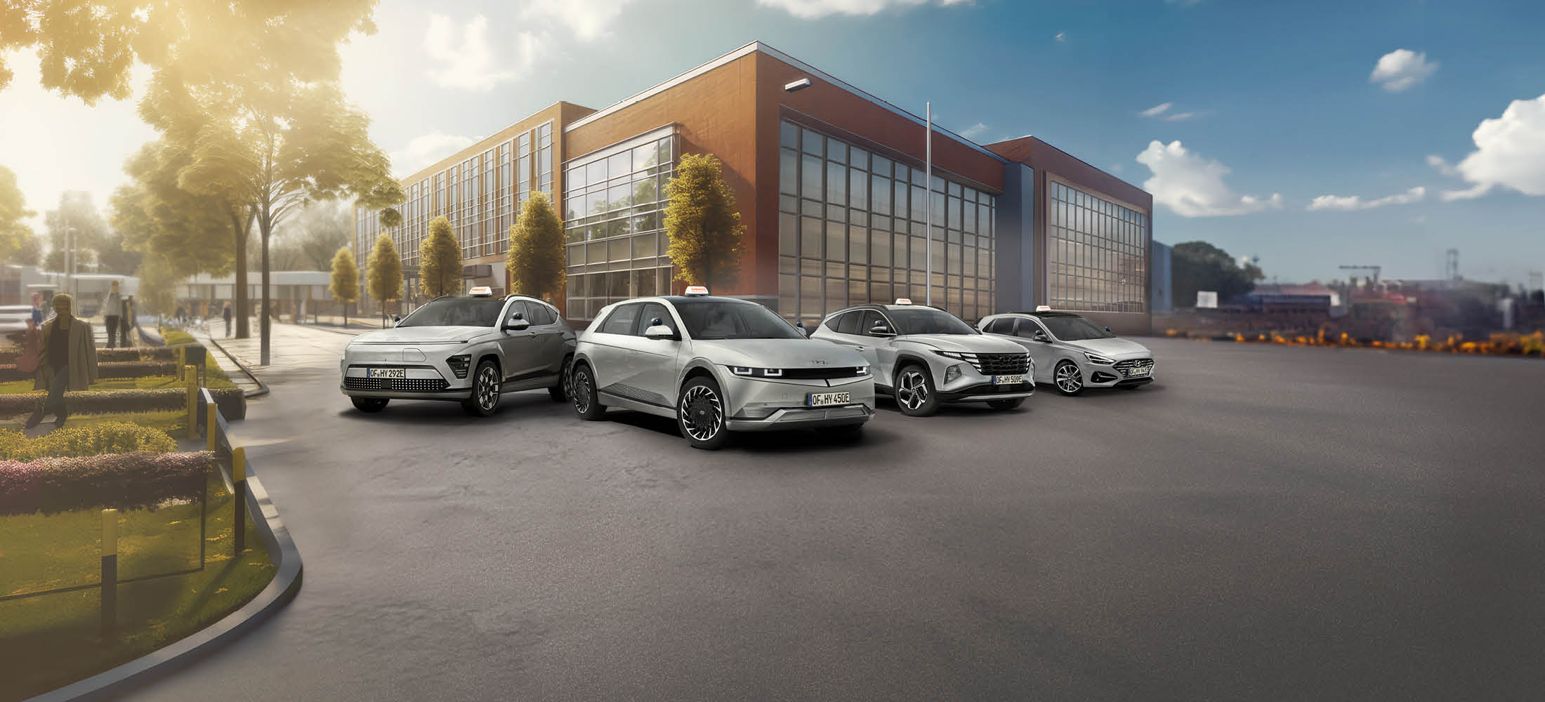 Vier moderne Fahrzeuge stehen nebeneinander auf einer großen, freien Fläche vor einem Schulgebäude. Alle Autos sind silberfarben und tragen Taxi-Schilder auf dem Dach.