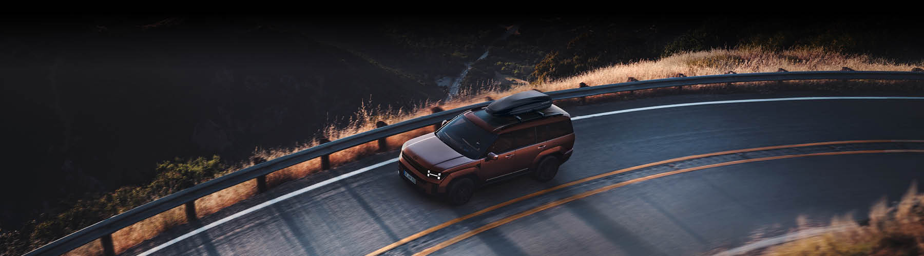 Ein brauner SUV mit Dachbox fährt auf einer kurvigen Bergstraße entlang eines Geländers, während Sonnenlicht das trockene Gras am Straßenrand beleuchtet.