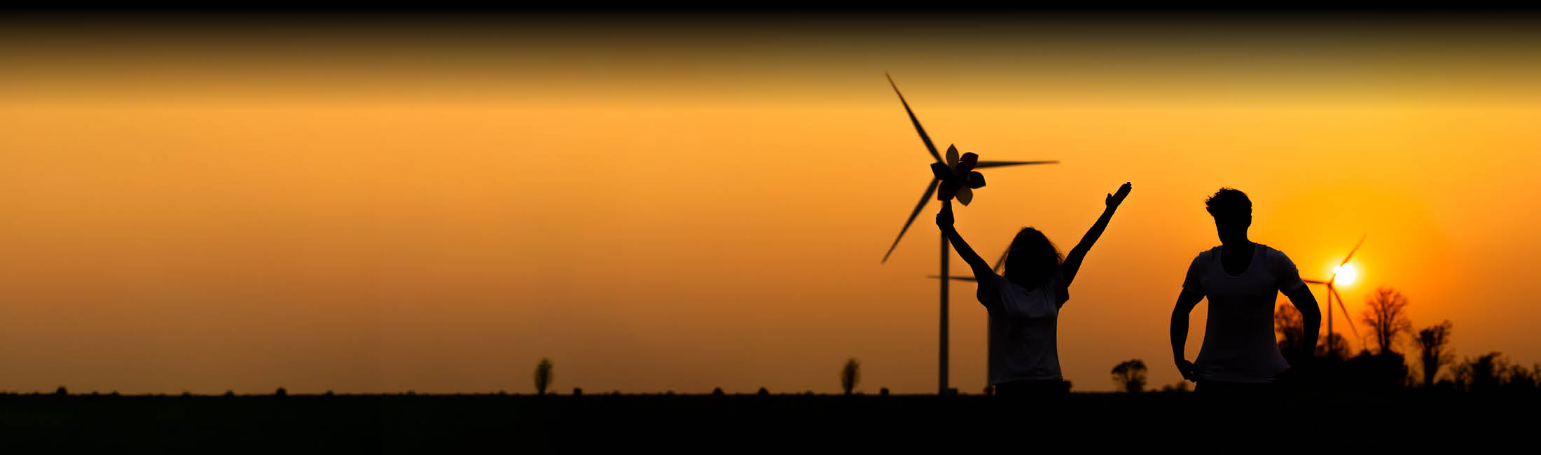 Zwei Silhouetten bei Sonnenuntergang auf einem Feld; im Hintergrund stehen Windräder. Eine Person hebt die Arme und hält ein kleines Windrad, die andere steht mit den Händen an der Hüfte. Der Himmel leuchtet orange.