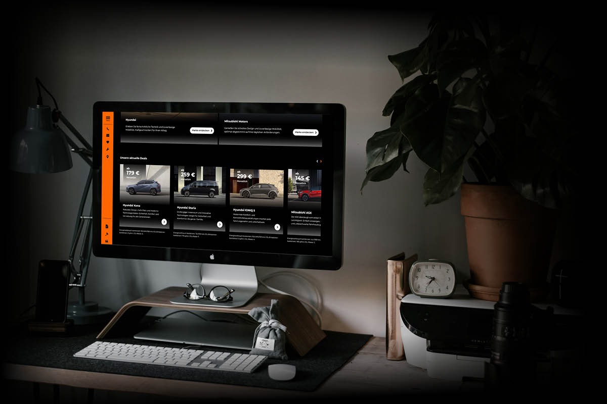 Auf einem Schreibtisch steht ein iMac-Computer, auf dem eine Website mit aktuellen Autoangeboten angezeigt wird. Es sind verschiedene Modelle und monatliche Preise sichtbar.
