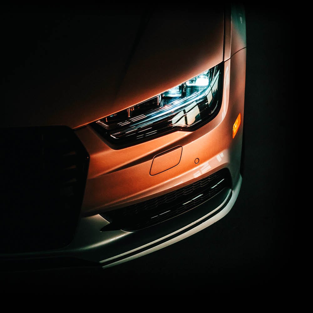 Das Bild zeigt die Front eines Autos im Dunkeln, wobei der Scheinwerfer und ein Teil des Kühlergrills sichtbar sind.