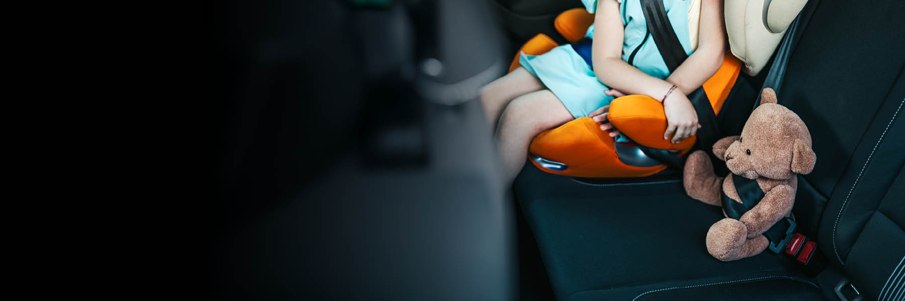 Ein Kind sitzt in einem Kindersitz auf dem Rücksitz eines Autos, daneben ist ein Teddybär mit dem Sicherheitsgurt angeschnallt.