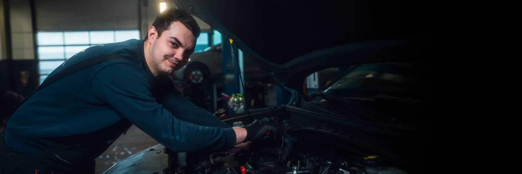 Eine Person arbeitet in einer Werkstatt an einem geöffneten Motorraum eines Autos.