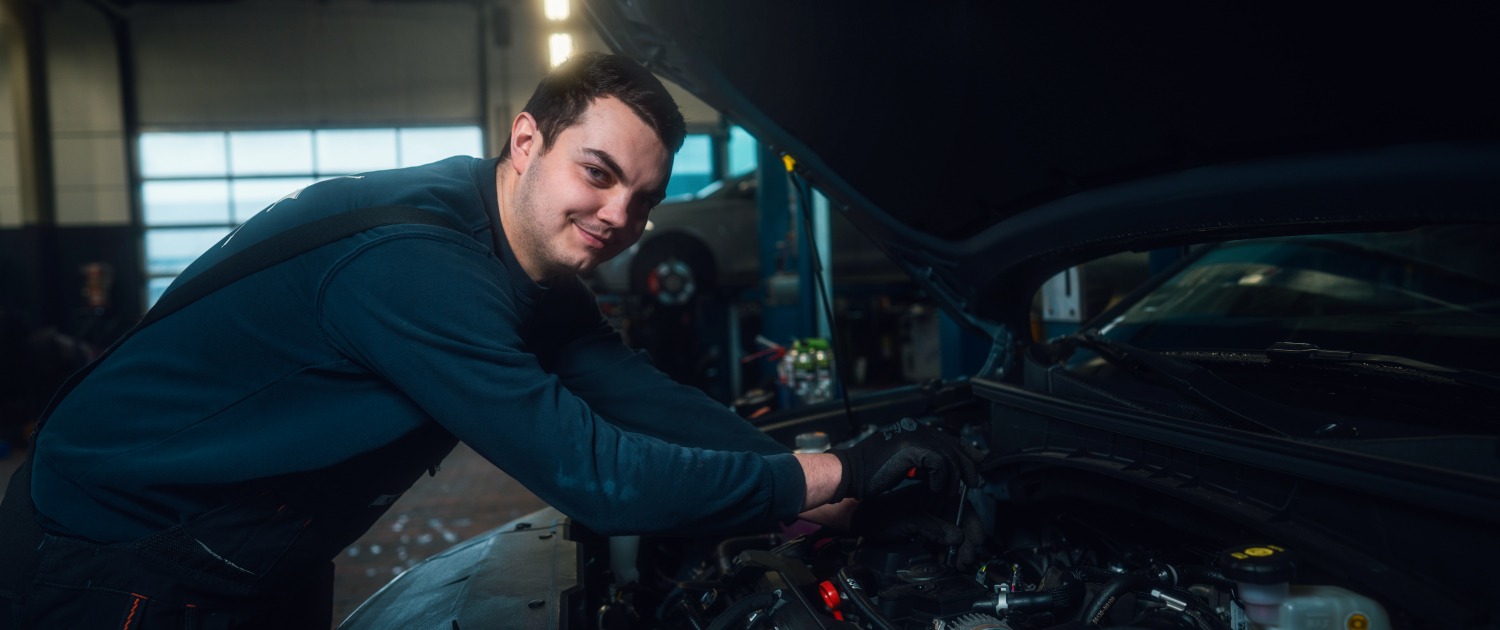 Eine Person arbeitet in einer Werkstatt an einem geöffneten Motorraum eines Autos.