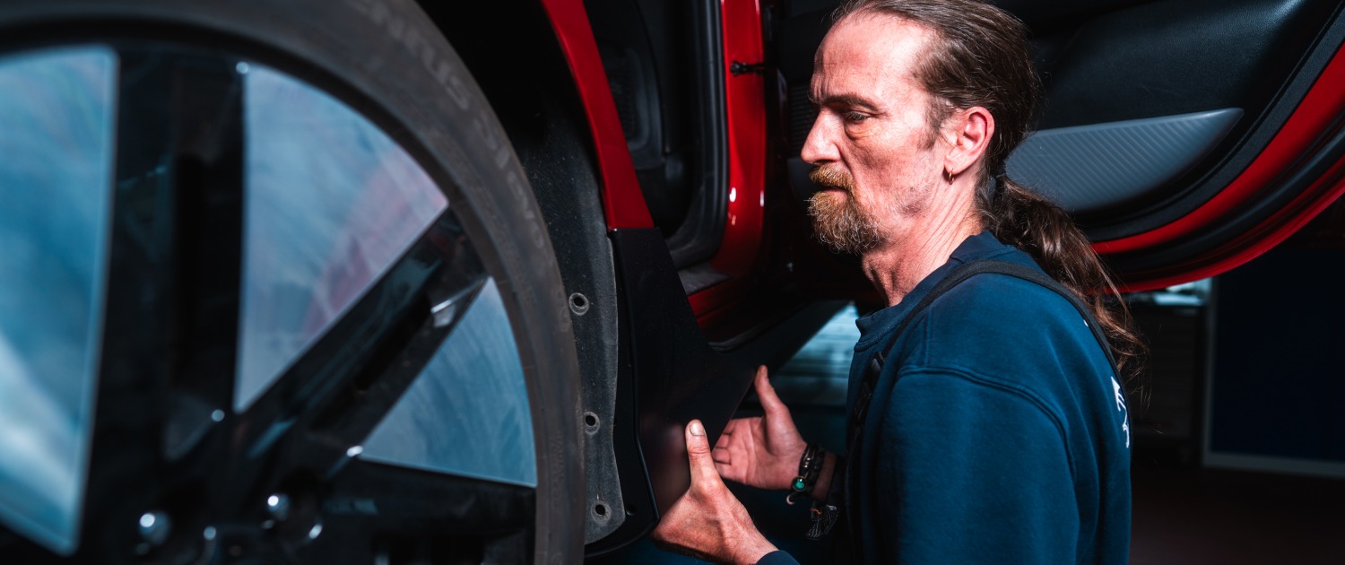 Eine Person montiert ein Bauteil, vermutlich einen Spritzschutz oder eine Verkleidung, im Bereich des Radkastens eines roten Fahrzeugs in einer Werkstatt.