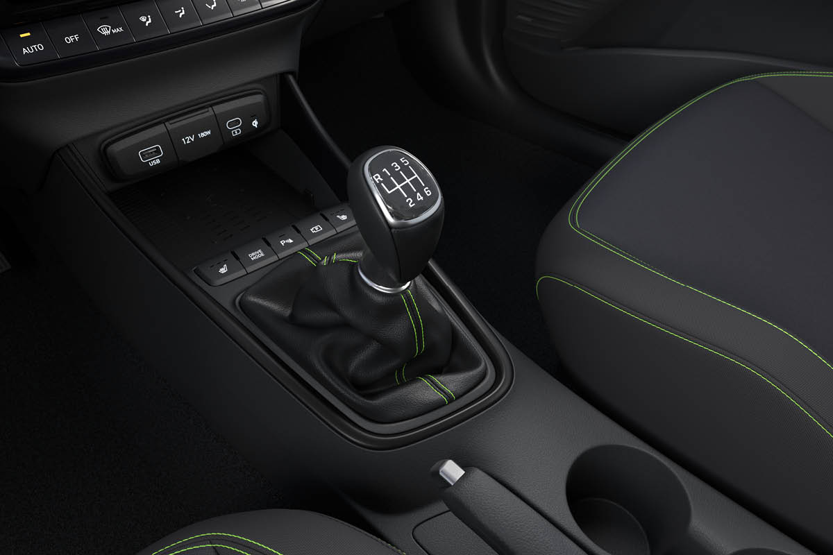 Nahaufnahme der Mittelkonsole eines Autos: ein 6‑Gang-Schaltknauf mit grünen Ziernähten, daneben Handbremse und Becherhalter. Vorne USB- und 12V-Anschluss sowie eine Ablage mit kabelloser Ladefläche, darüber Klimabedienelemente.