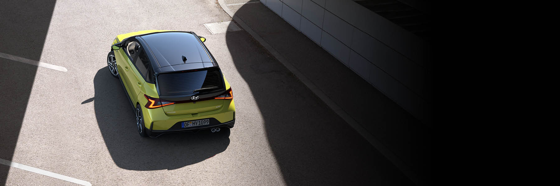 Schräg-obenansicht eines gelbgrünen Hatchbacks mit schwarzem Dach: markante LED‑Rückleuchten, Doppel-Endrohr und sportliche Felgen, fährt durch eine teils schattige Stadtpassage.