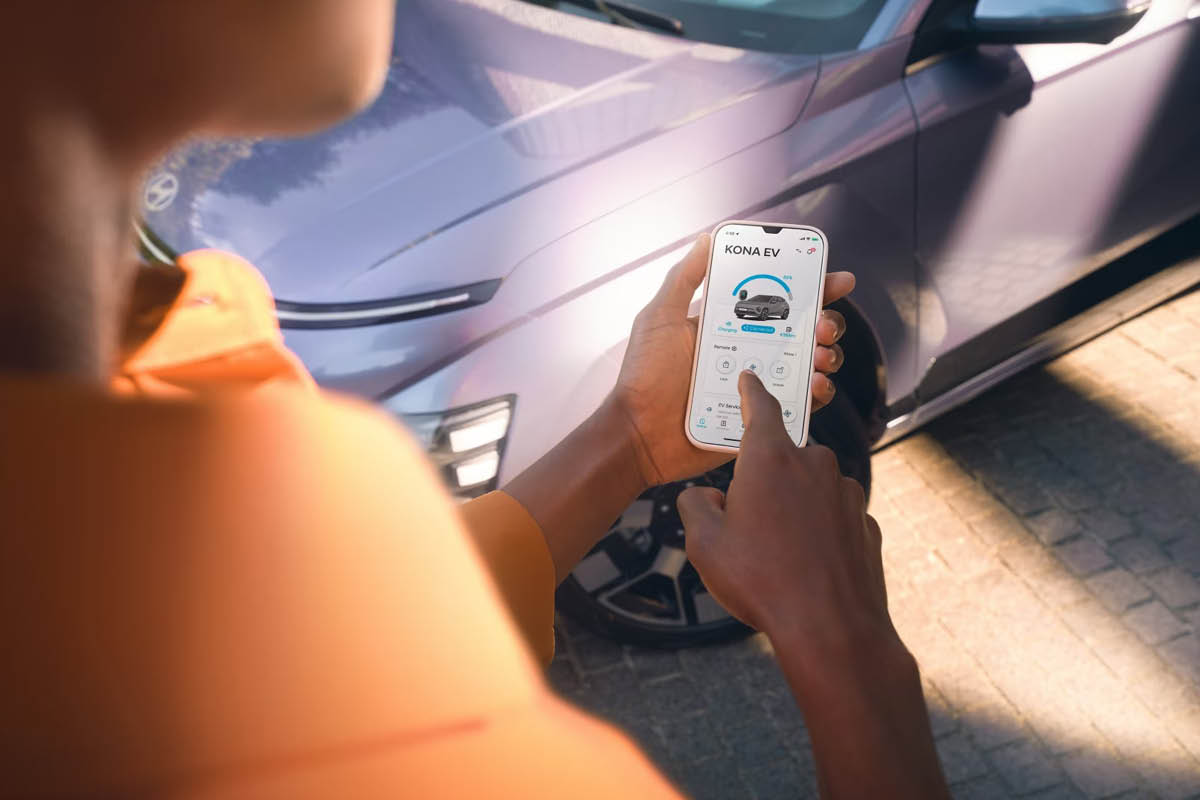 Eine Person steht vor einem modernen Auto und steuert über eine Smartphone-App mit der Anzeige "KONA EV" verschiedene Funktionen des Fahrzeugs.