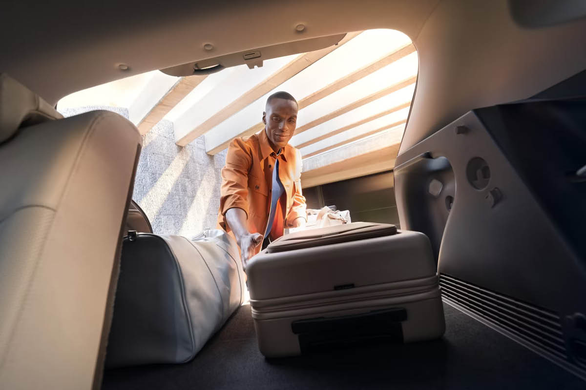Das Bild zeigt, wie eine Person Koffer und Gepäck in den Kofferraum eines Autos lädt. Die Szene wird aus dem Inneren des Autos fotografiert.