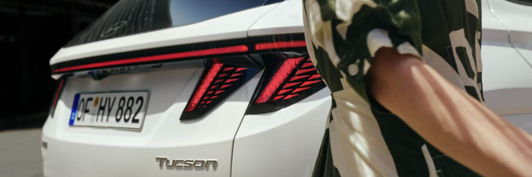 Heckansicht eines weißen SUVs mit durchgehender roter LED-Lichtleiste und markanten, dreiecksmusterartigen Rückleuchten; „TUCSON“-Schriftzug und EU‑Kennzeichen sichtbar, rechts läuft eine Person vorbei.